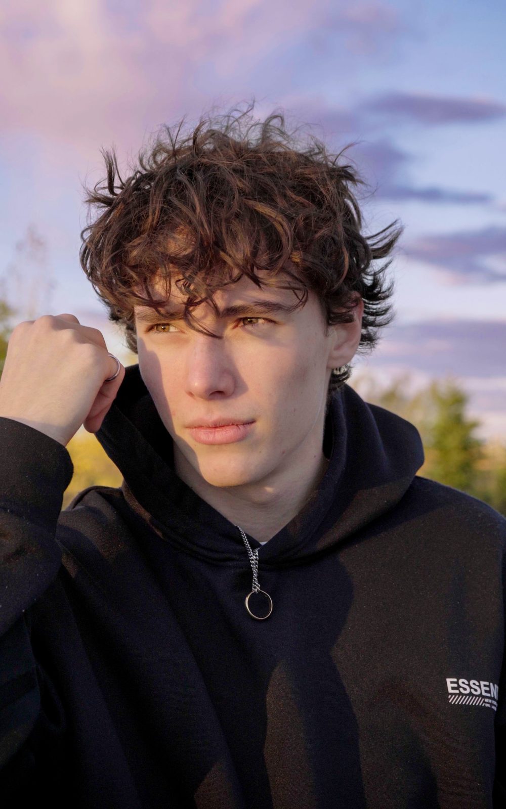 Portrait d'un jeune homme aux cheveux bouclés en chandail à capuchon, sous un ciel rose et bleu