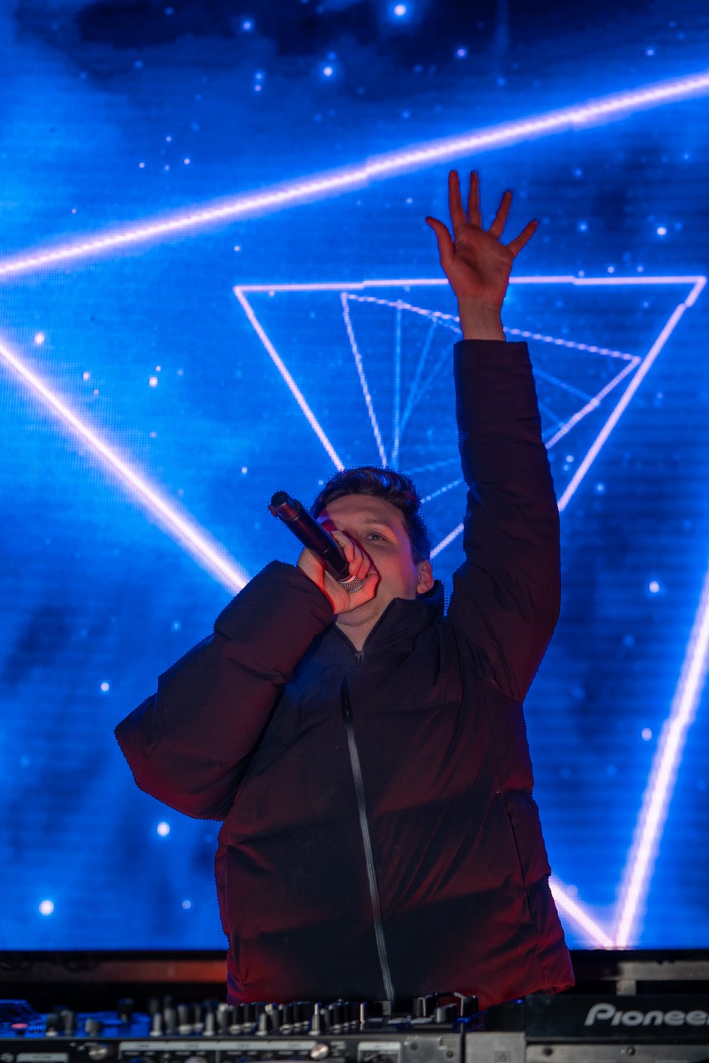 Le DJ Mike Demero chantant au micro, bras levé, devant un écran bleu aux motifs de triangles lumineux