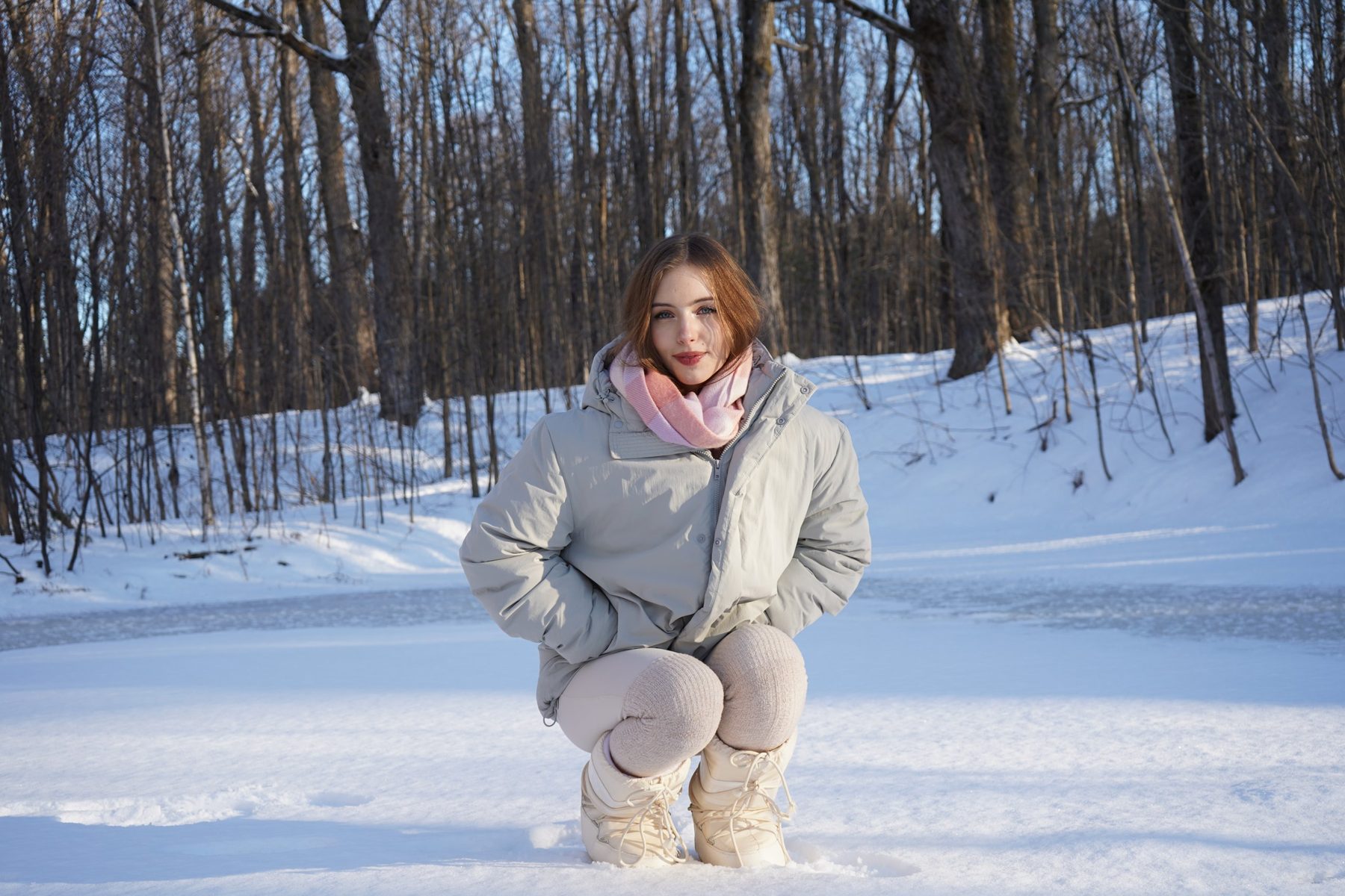 Jeune femme accroupie dans la neige, en manteau gris et foulard rose, devant une forêt d'hiver