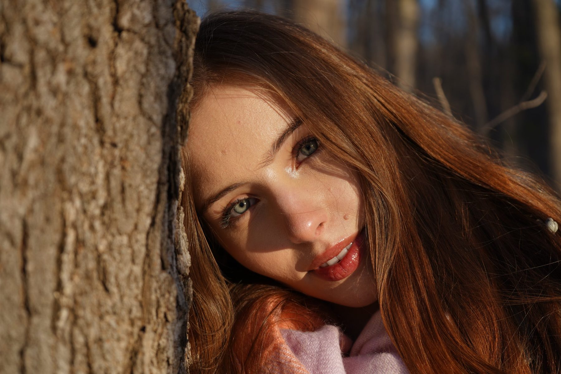 Gros plan d'une jeune femme aux cheveux roux appuyée contre un tronc d'arbre, au soleil