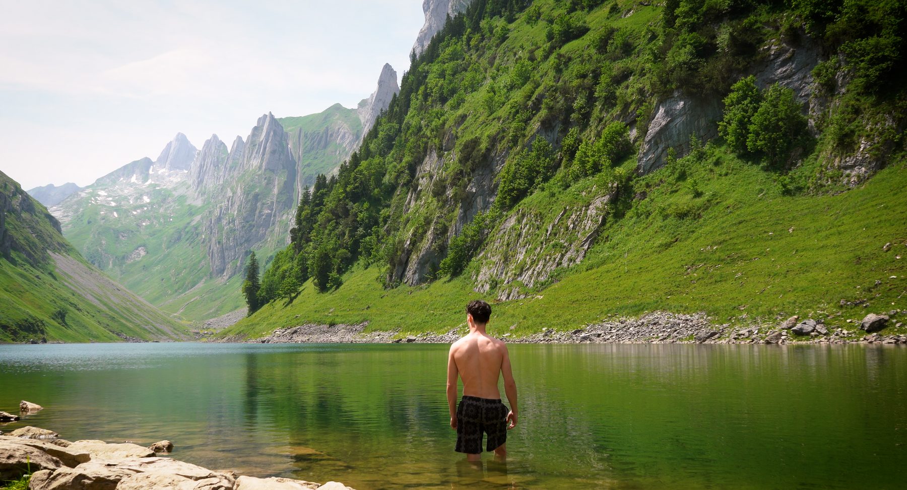Homme de dos, debout dans un lac alpin vert entouré de falaises et de montagnes