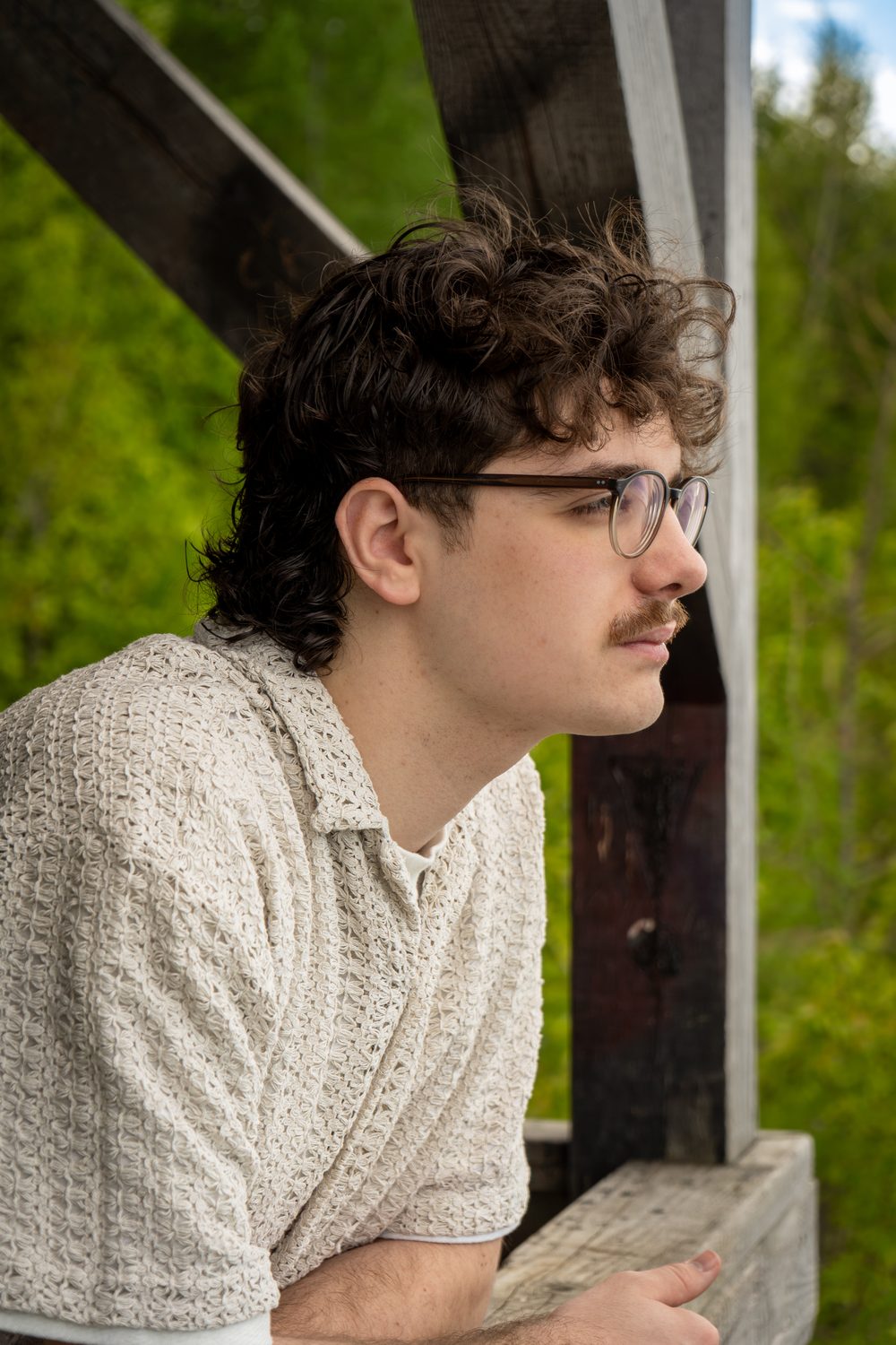 Portrait de profil d'un jeune homme aux cheveux bouclés et aux lunettes, accoudé à une rambarde en bois devant la forêt