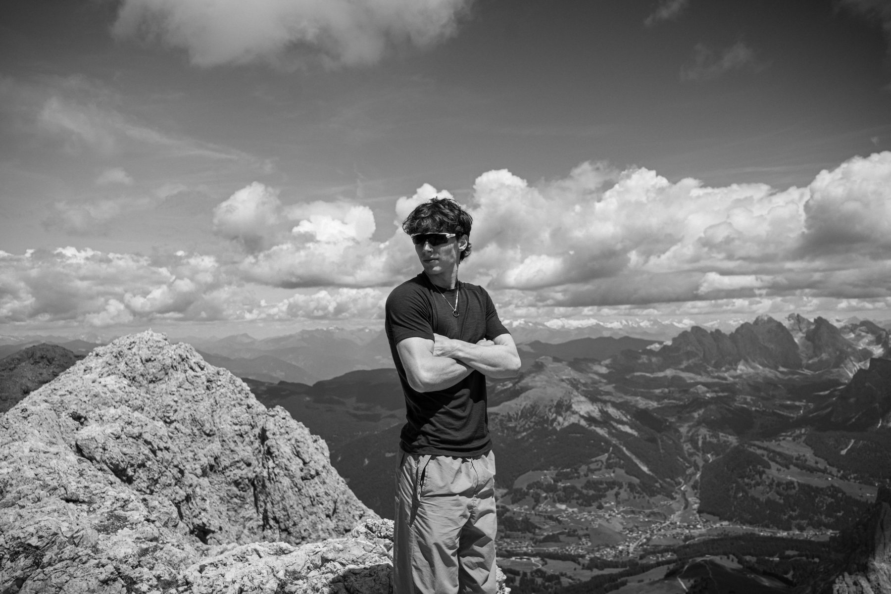 Portrait en noir et blanc d'un jeune homme aux lunettes de soleil, bras croisés au sommet d'une montagne